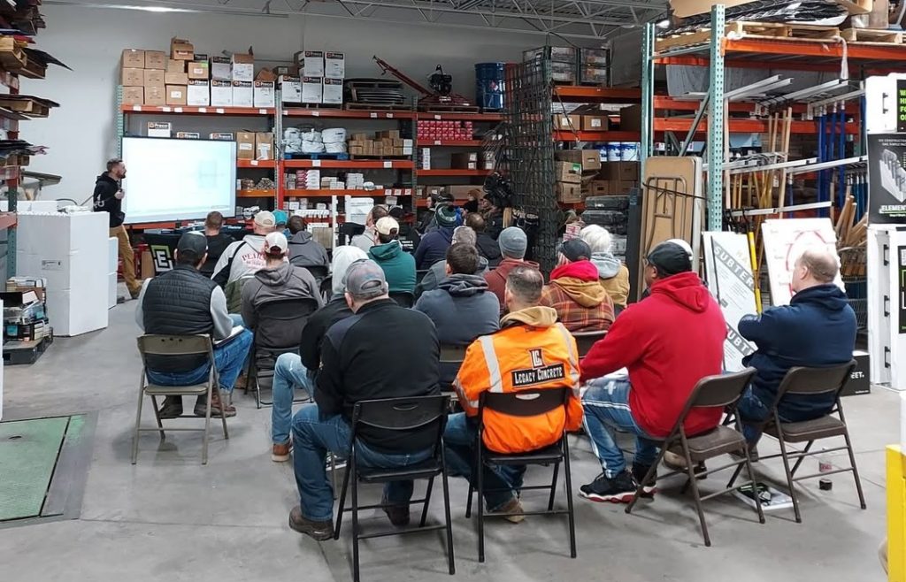 Group of people seated on folding chairs in a warehouse, watching a presenter give a talk with a projection screen at the front.